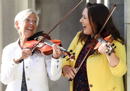 Two women play the fiddle together in the sunshine